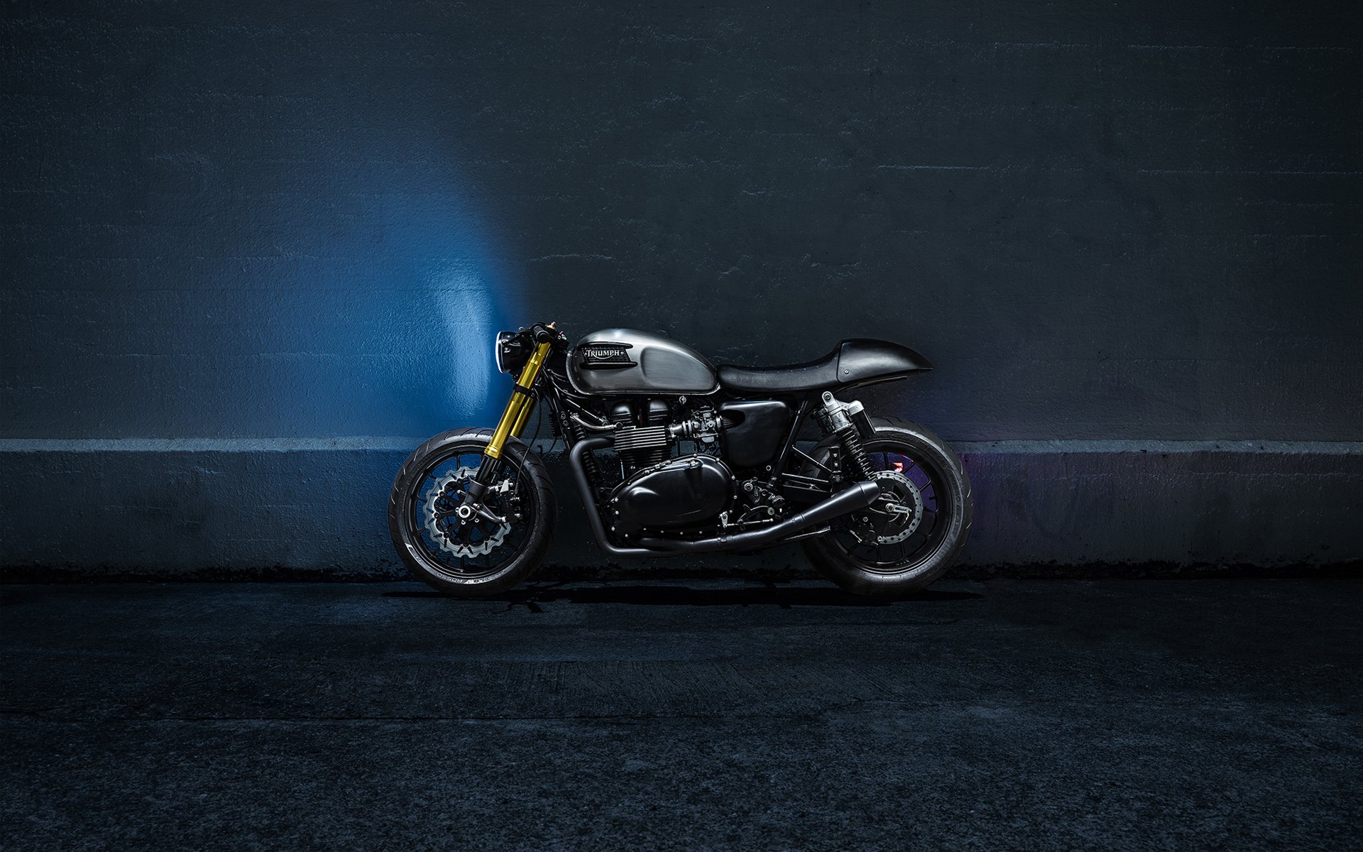 Side view of a black Triumph Bonneville The Bullitt motorcycle against a moody textured wall, HD PC desktop wallpaper background.