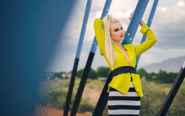 A blonde Australian rapper and singer poses confidently outdoors, dressed in a vibrant yellow jacket and striped skirt, against a backdrop of blue structures and a scenic landscape.