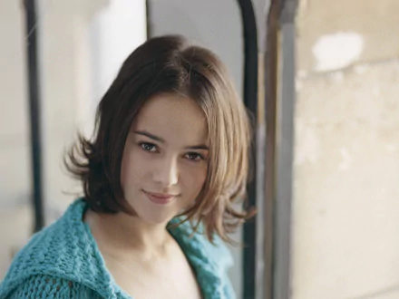 HD desktop wallpaper featuring Alizée with a soft smile, set against a blurred indoor background, highlighting her natural beauty and musical persona.