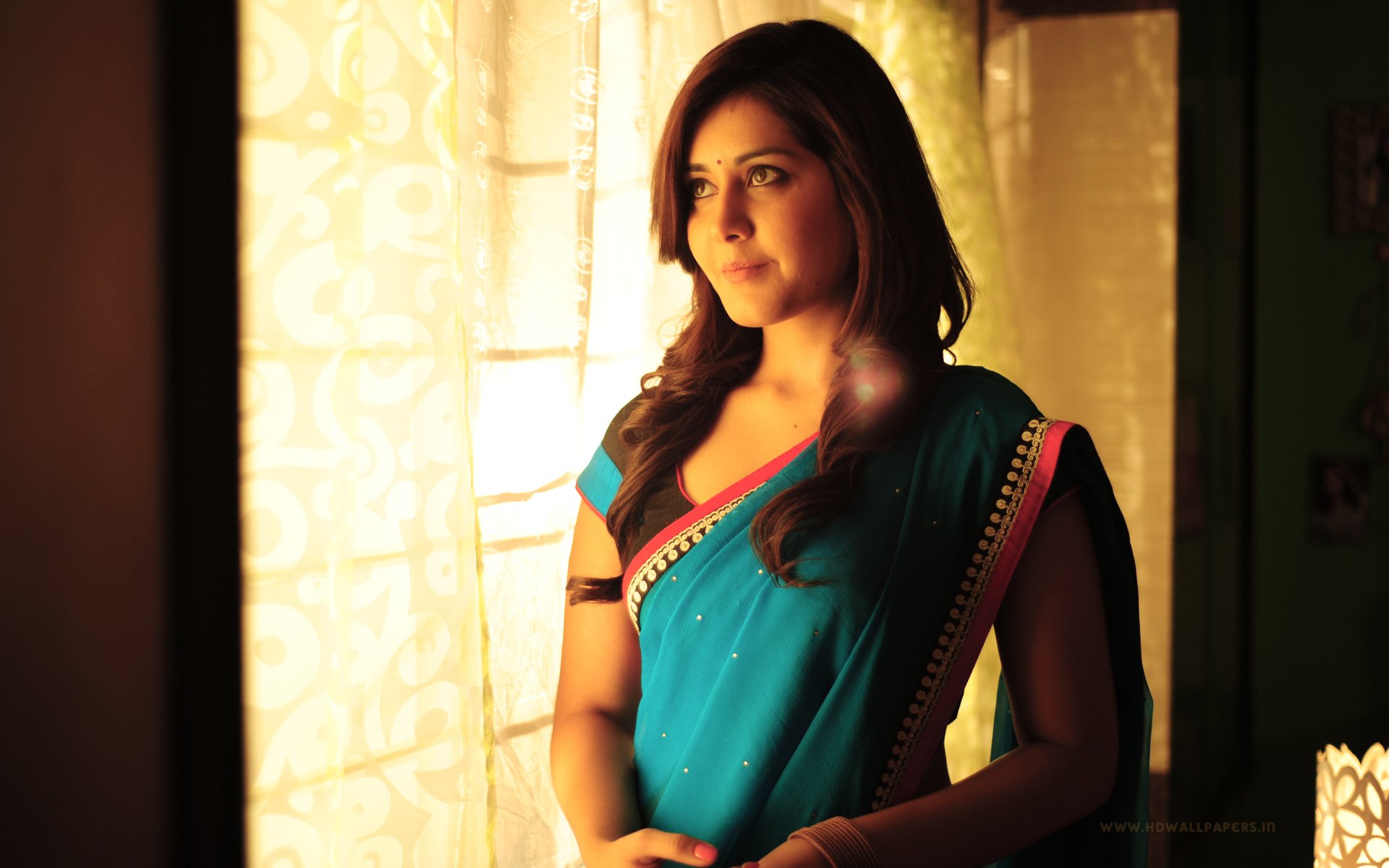 A stunning HD desktop wallpaper featuring celebrity Rashi Khanna in a blue saree, standing by a sunlit window, exuding elegance and grace as she gazes thoughtfully into the distance.