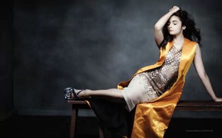 A HD desktop wallpaper featuring Alia Bhatt, elegantly posed with one leg draped over a bench, wearing a stylish outfit complemented by an eye-catching orange robe.