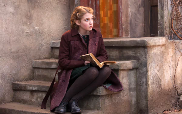 Sophie Nélisse as Liesel in the movie The Book Thief, sitting on stone steps holding an open book, with a rustic wall and doorway in the background.