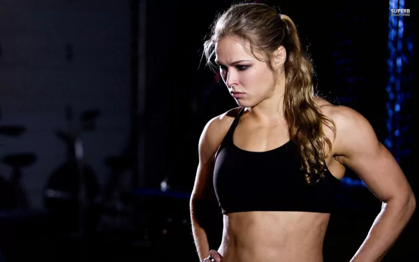 A focused Ronda Rousey stands in a gym, wearing a black sports top, showcasing her athleticism and determination. This HD image serves as an inspiring sports-themed desktop background.