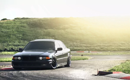 Black BMW E38 parked on a sunlit track with grassy hills; HD PC desktop wallpaper background, warm light and low-angle perspective.