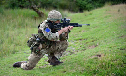 HD desktop wallpaper of a soldier in full military gear aiming a rifle while kneeling on grassy terrain.