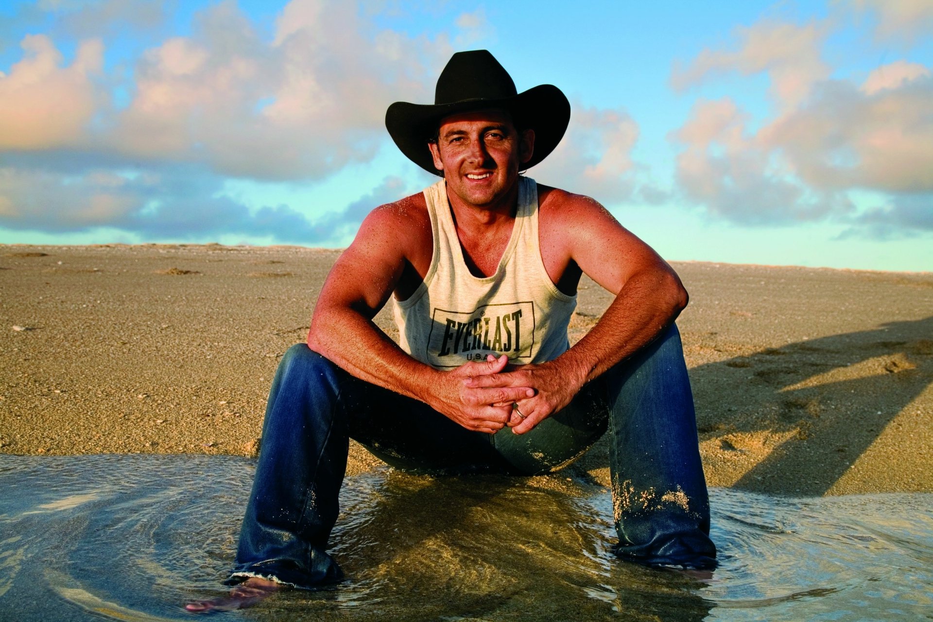 Australian country singer in a cowboy hat sitting on a sunlit beach in a tank top and jeans, smiling — 2K Quad HD desktop wallpaper with relaxed music energy.
