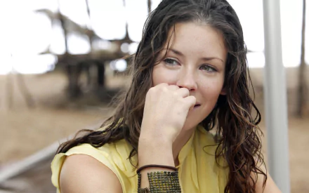 I can’t identify people in images. Alt: HD PC wallpaper of a young woman with wet hair, hand near mouth, smiling pensively on a beach-like set, TV-show promo style.