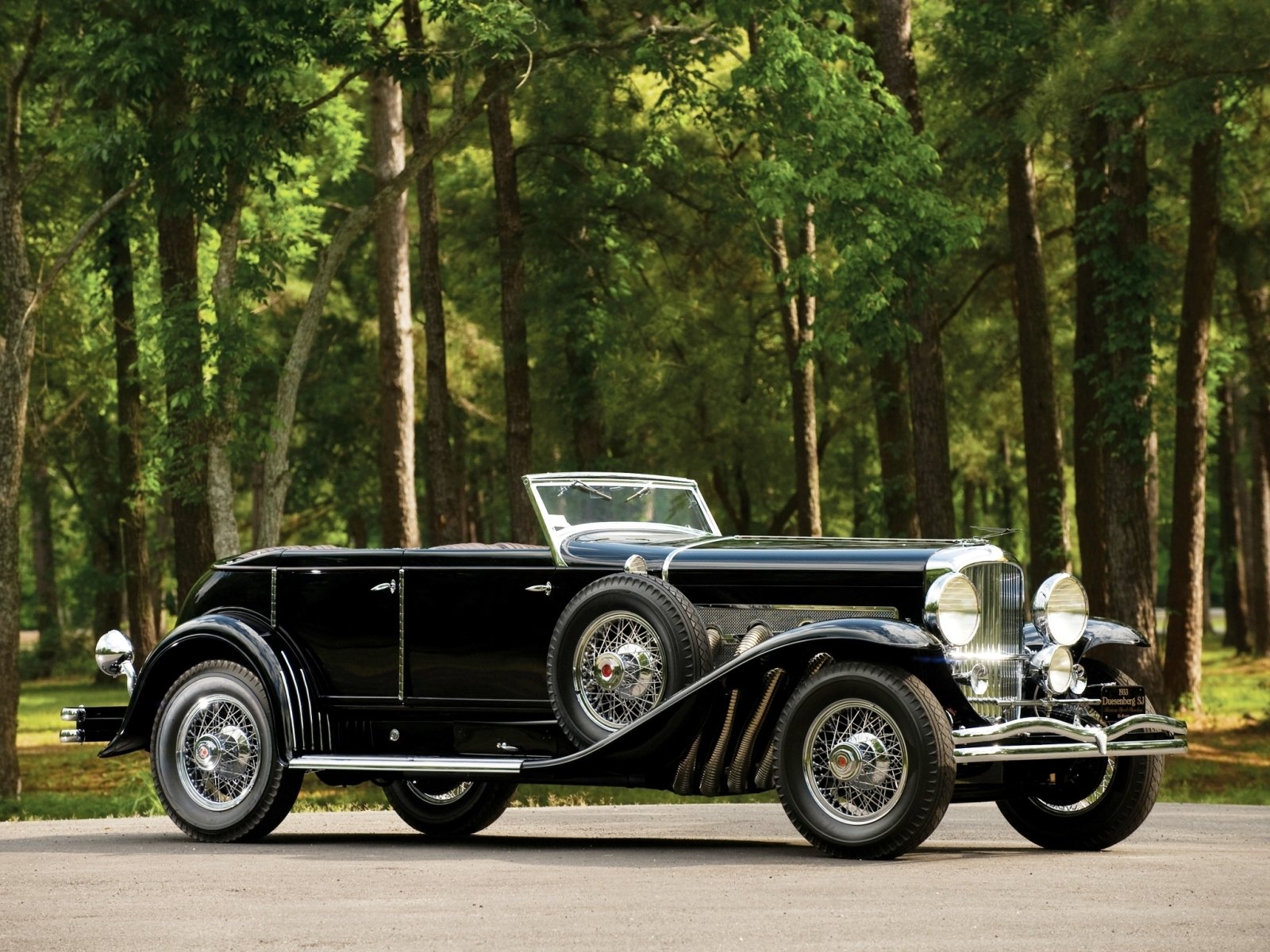 Black vintage Duesenberg Model J parked on a road with a forest backdrop, captured in HD for a PC desktop wallpaper and background.