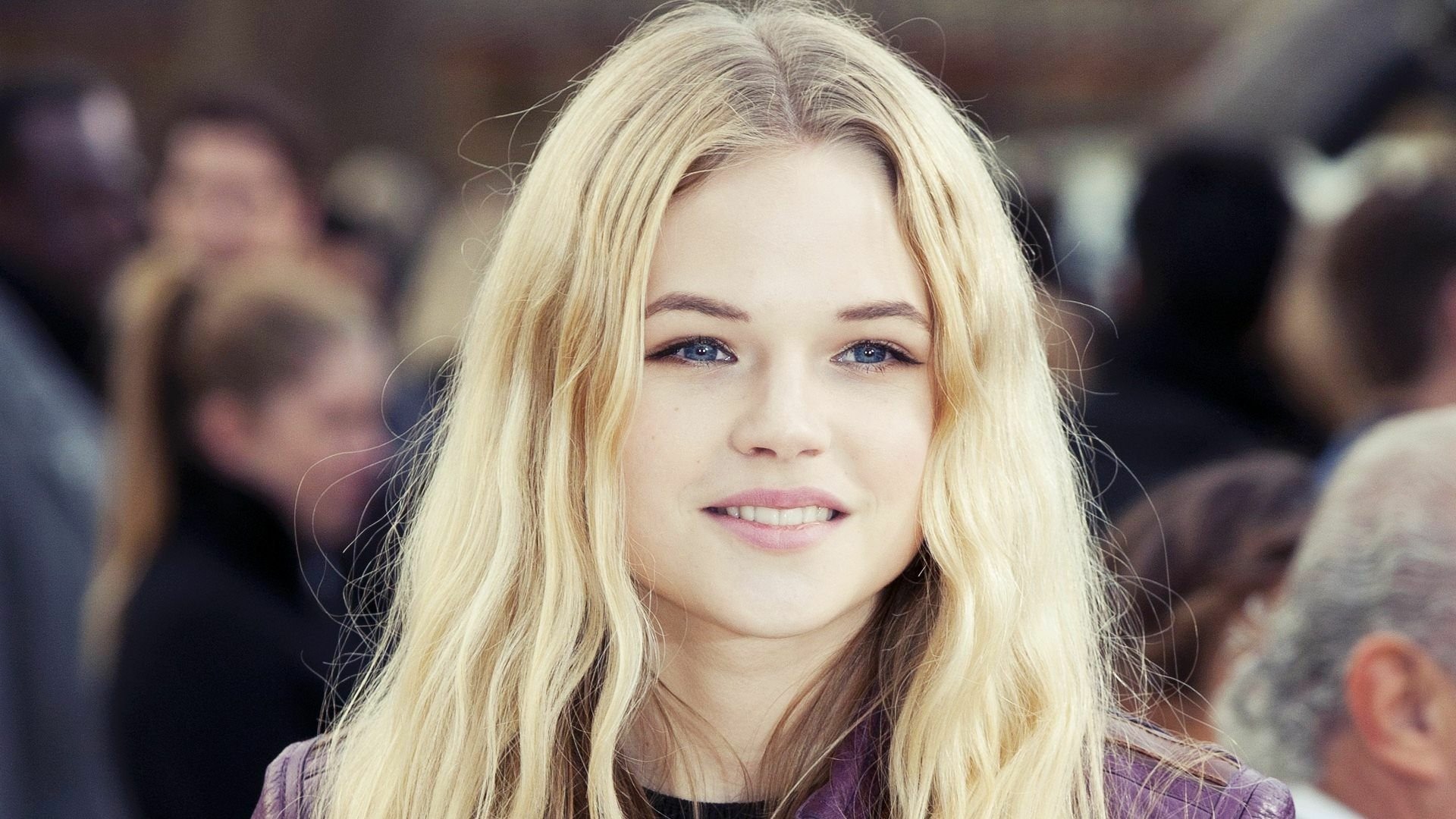 HD PC desktop wallpaper featuring a close-up portrait of woman Gabriella Wilde with long blonde hair and a soft smile, set against a blurred crowd background.