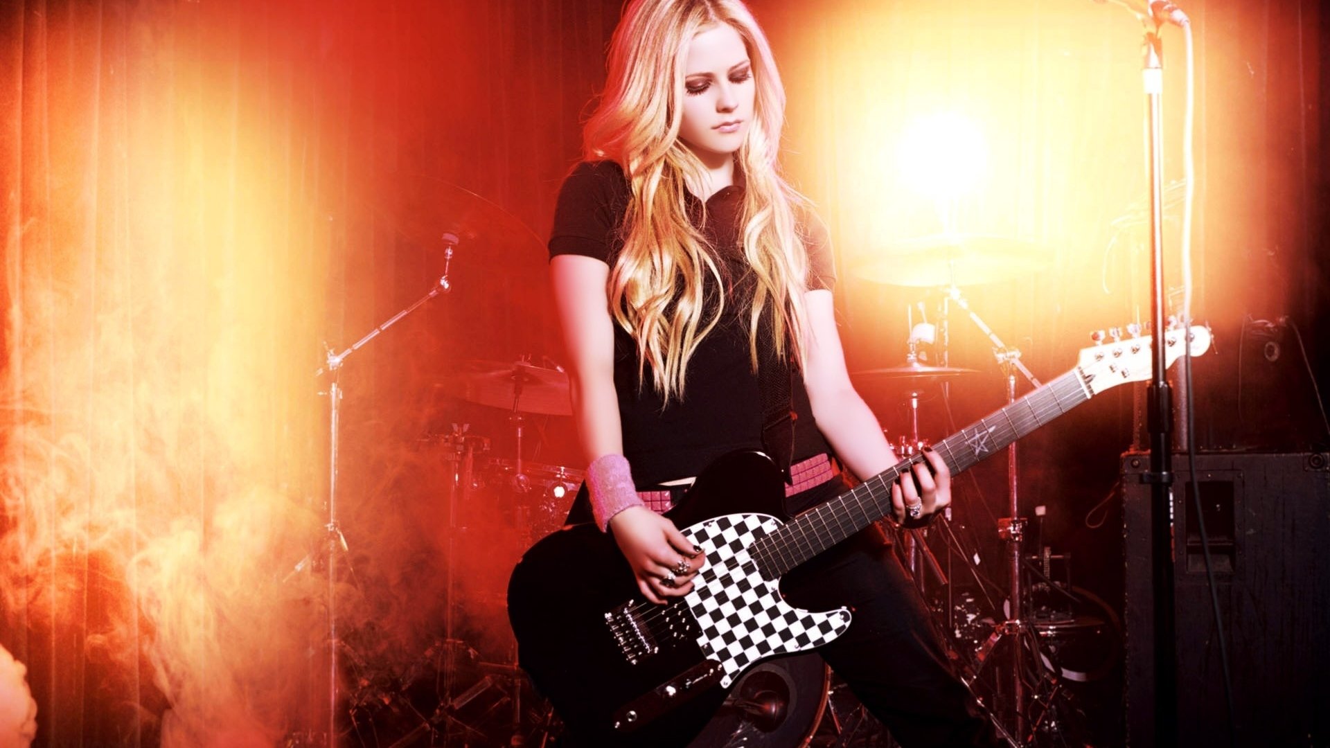 HD desktop wallpaper of Avril Lavigne playing a black and white checkered electric guitar on stage, surrounded by warm orange lighting and musical equipment.