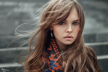 HD desktop wallpaper featuring Anastasiya Scheglova with a contemplative expression, standing against a blurred, gray background. Her hair flows with the wind, and she wears a patterned scarf.