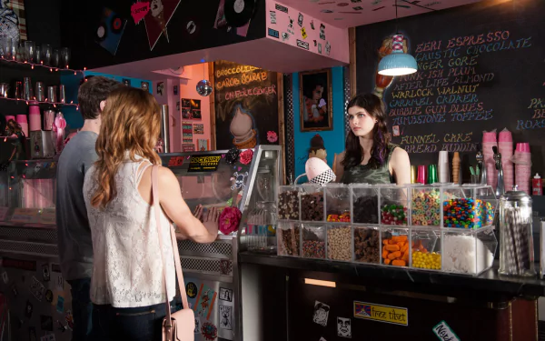 A scene from the movie Burying the Ex, featuring a vibrant ice cream shop with a customer at the counter and a server behind it, showcasing colorful candy options.