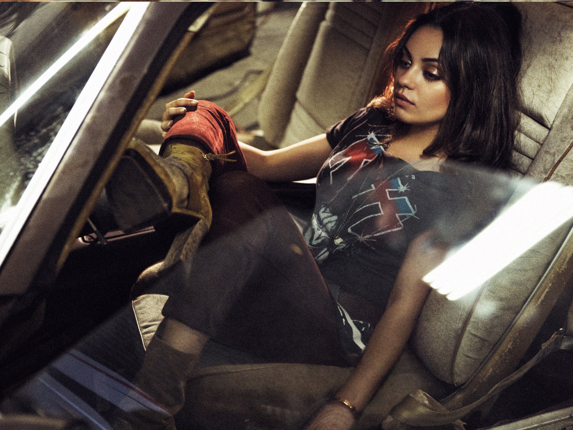 HD desktop wallpaper featuring celebrity Mila Kunis sitting inside a car, wearing a graphic t-shirt and boots, captured in a moody, cinematic style.
