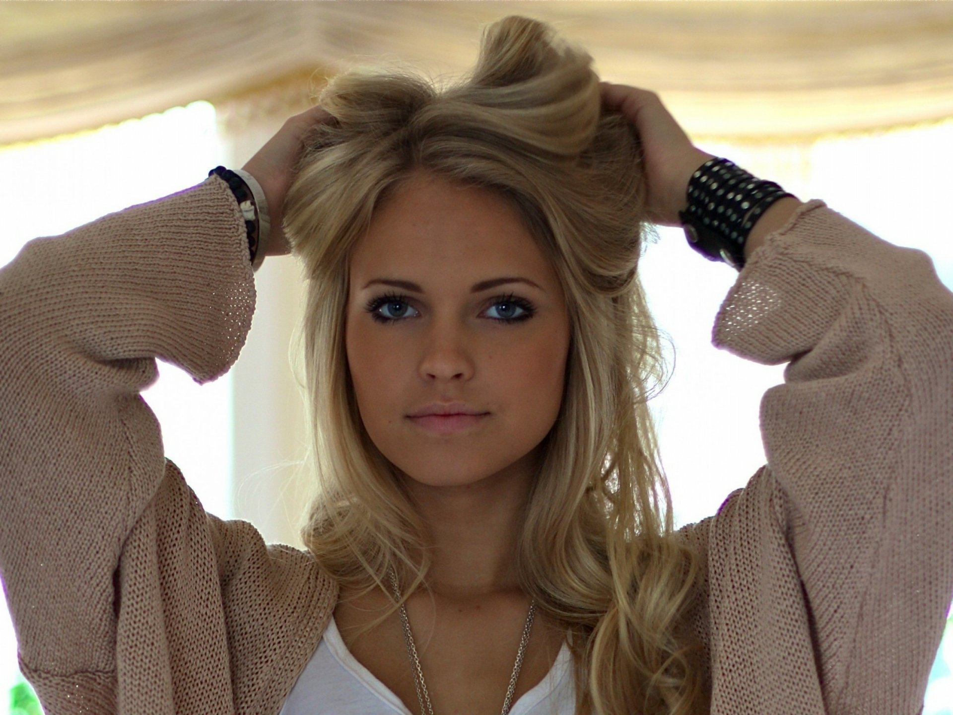 HD desktop wallpaper featuring a woman tagged as Emilie Marie Nereng, with a serene expression, holding her blonde hair up, in a softly lit, cozy indoor setting.