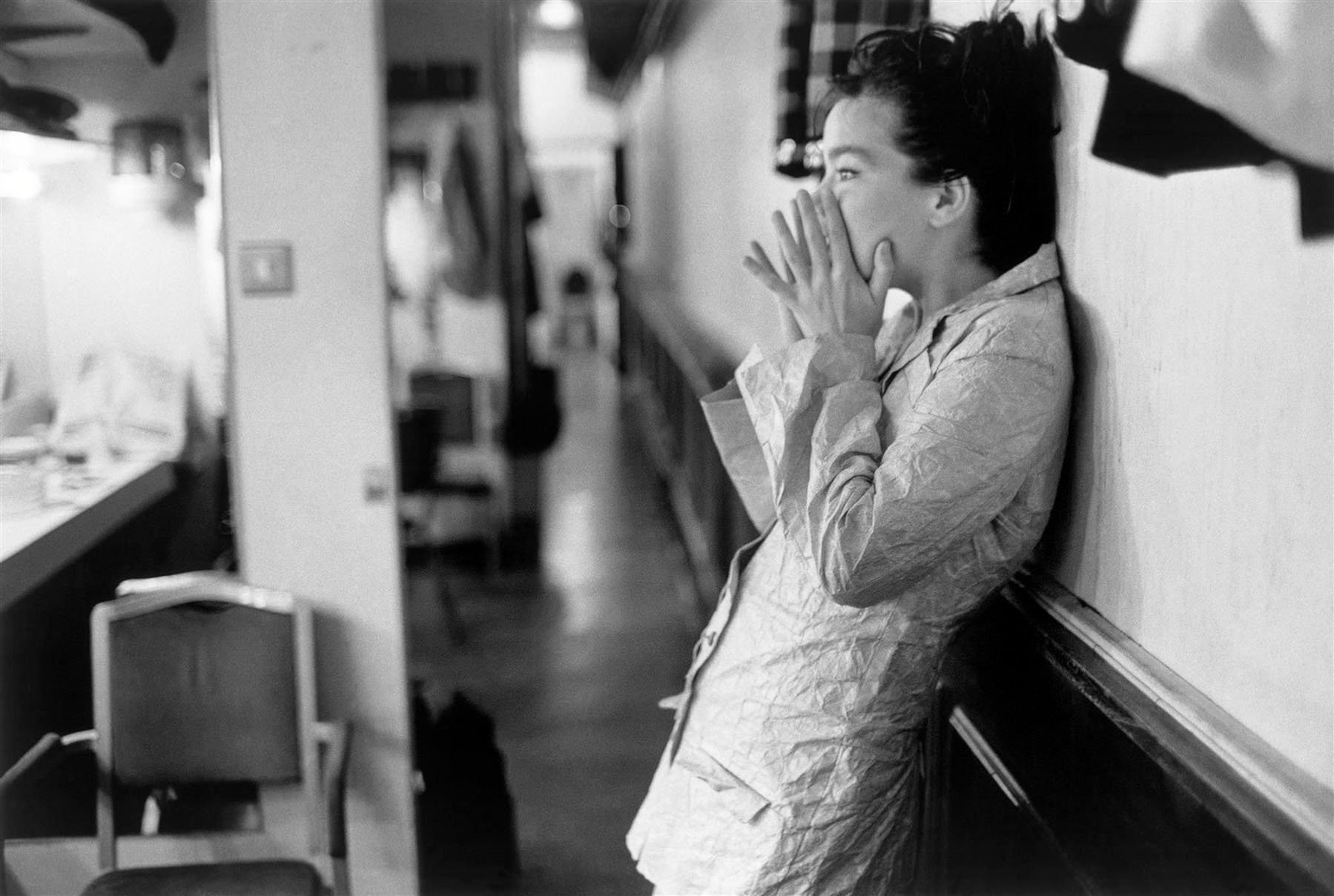 Black-and-white HD desktop wallpaper of Björk in a candid moment, leaning against a wall in a backstage or dressing room setting, capturing a music-related vibe.