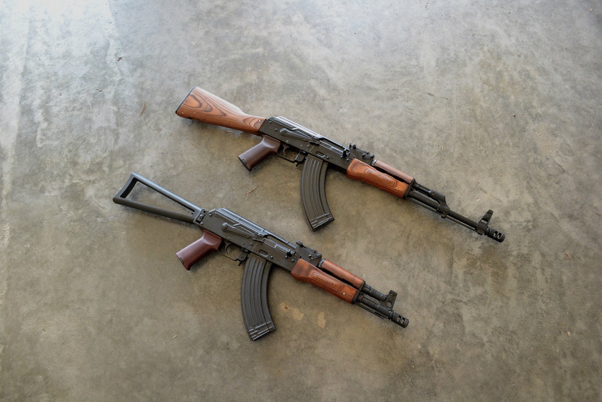 Two AK-47 rifles are displayed on a concrete surface, showcasing their distinct design and craftsmanship, serving as a striking HD desktop wallpaper and background.