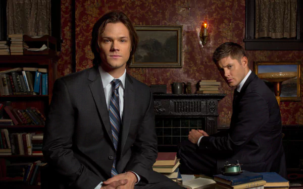 Two characters from the TV show Supernatural pose in a vintage room filled with books. One sits confidently in a suit, while the other looks contemplative in the background.