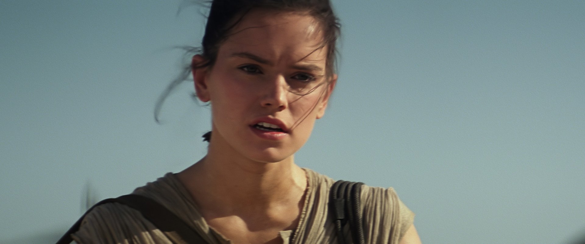 Rey, portrayed by Daisy Ridley, from Star Wars Episode VII: The Force Awakens, gazes intensely against a blue sky in this high-definition desktop wallpaper.