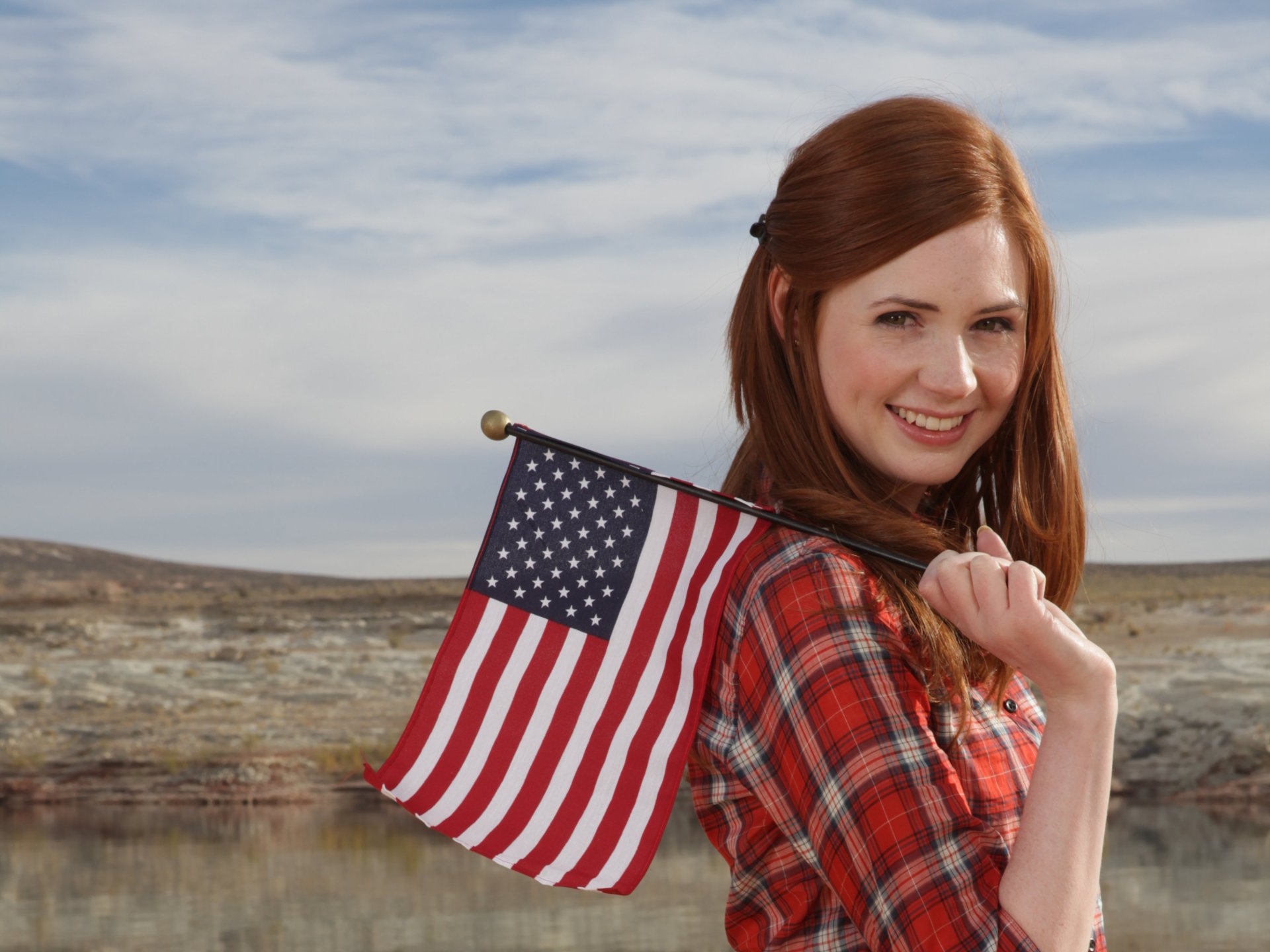 2K Quad HD PC desktop wallpaper of a smiling red-haired woman in a plaid shirt holding a U.S. flag against a wide, cloudy outdoor landscape.