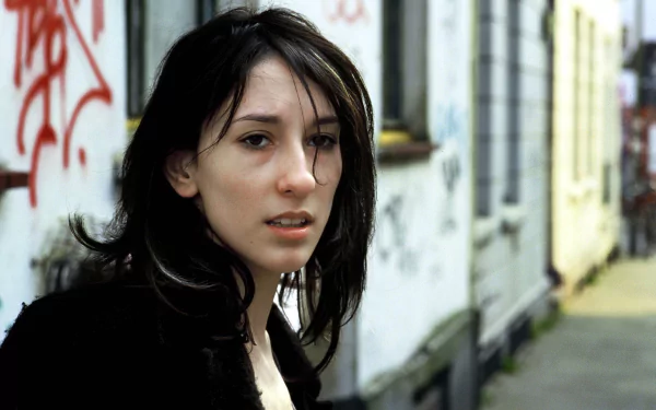 A HD desktop wallpaper featuring actress Sibel Kekilli, standing in an urban setting with graffiti-covered walls, showcasing a contemplative expression and striking features.