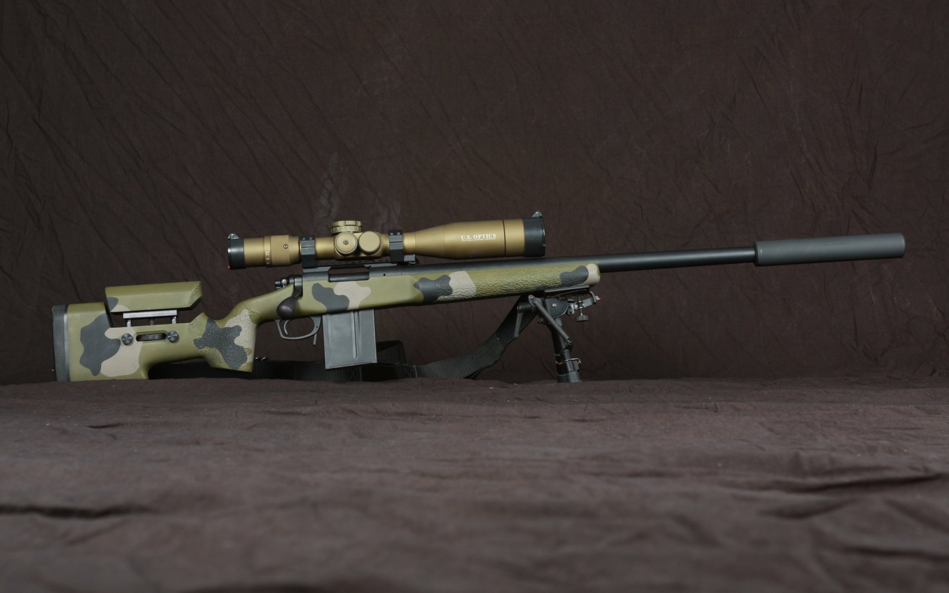 HD desktop wallpaper of a man-made rifle with a camouflage stock, equipped with a scope and suppressor, resting on a bipod against a dark background.