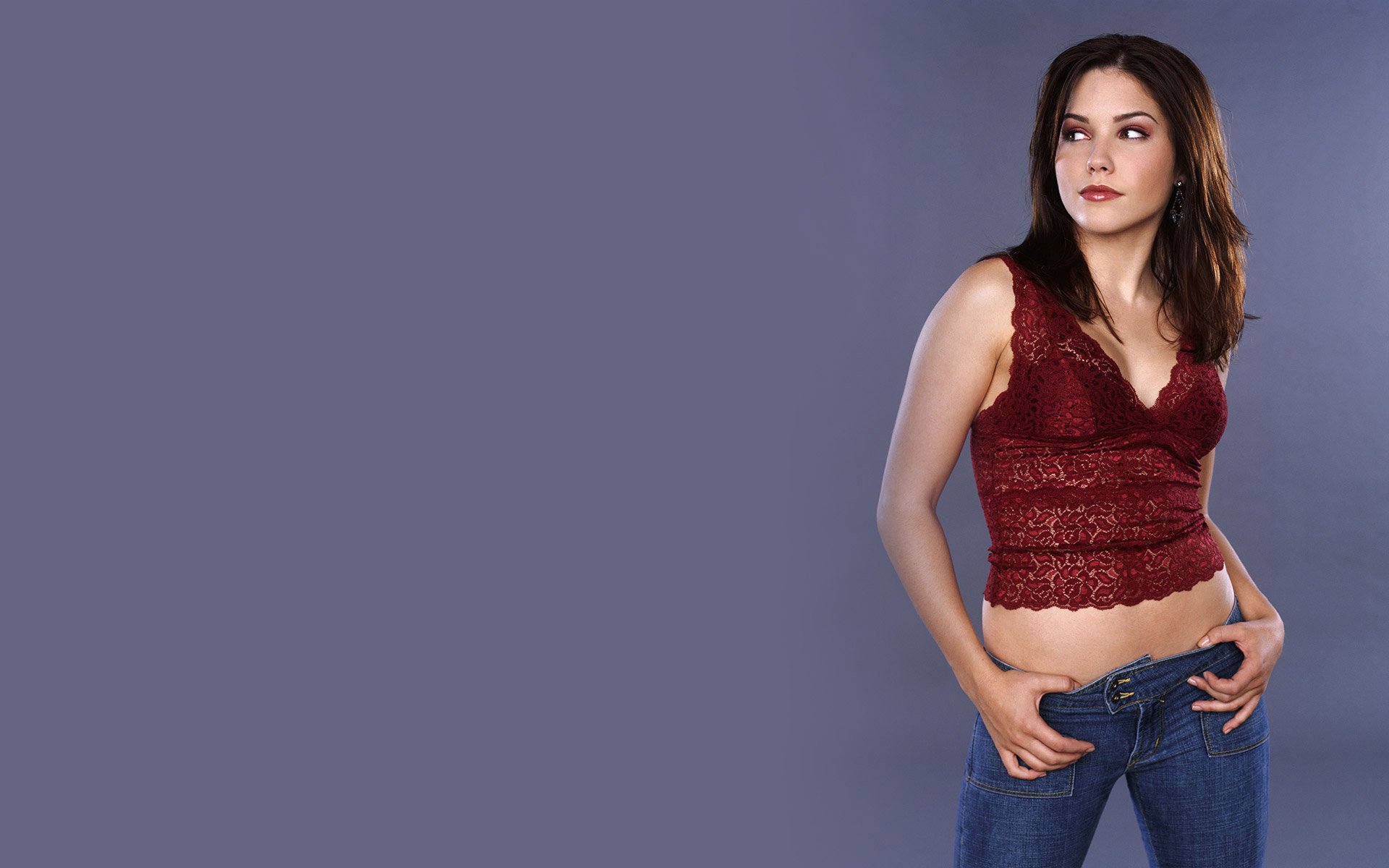 HD PC desktop wallpaper of a female celebrity in a burgundy lace camisole and low-rise jeans, posing against a soft gray background with hands at her waistband.