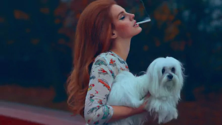 A woman with long red hair holds a small white dog while smoking a cigarette, set against a softly blurred background, capturing a moody ambiance reminiscent of Lana Del Rey.