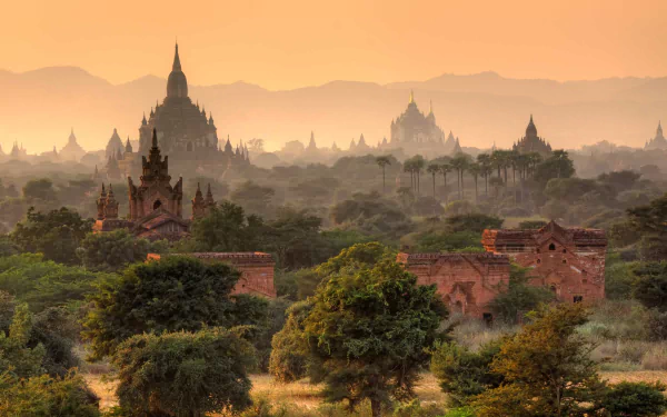 A stunning view of Bagan, Myanmar, showcasing its ancient temples silhouetted against a warm sunset, surrounded by lush greenery and distant mountains. An inspiring HD wallpaper.