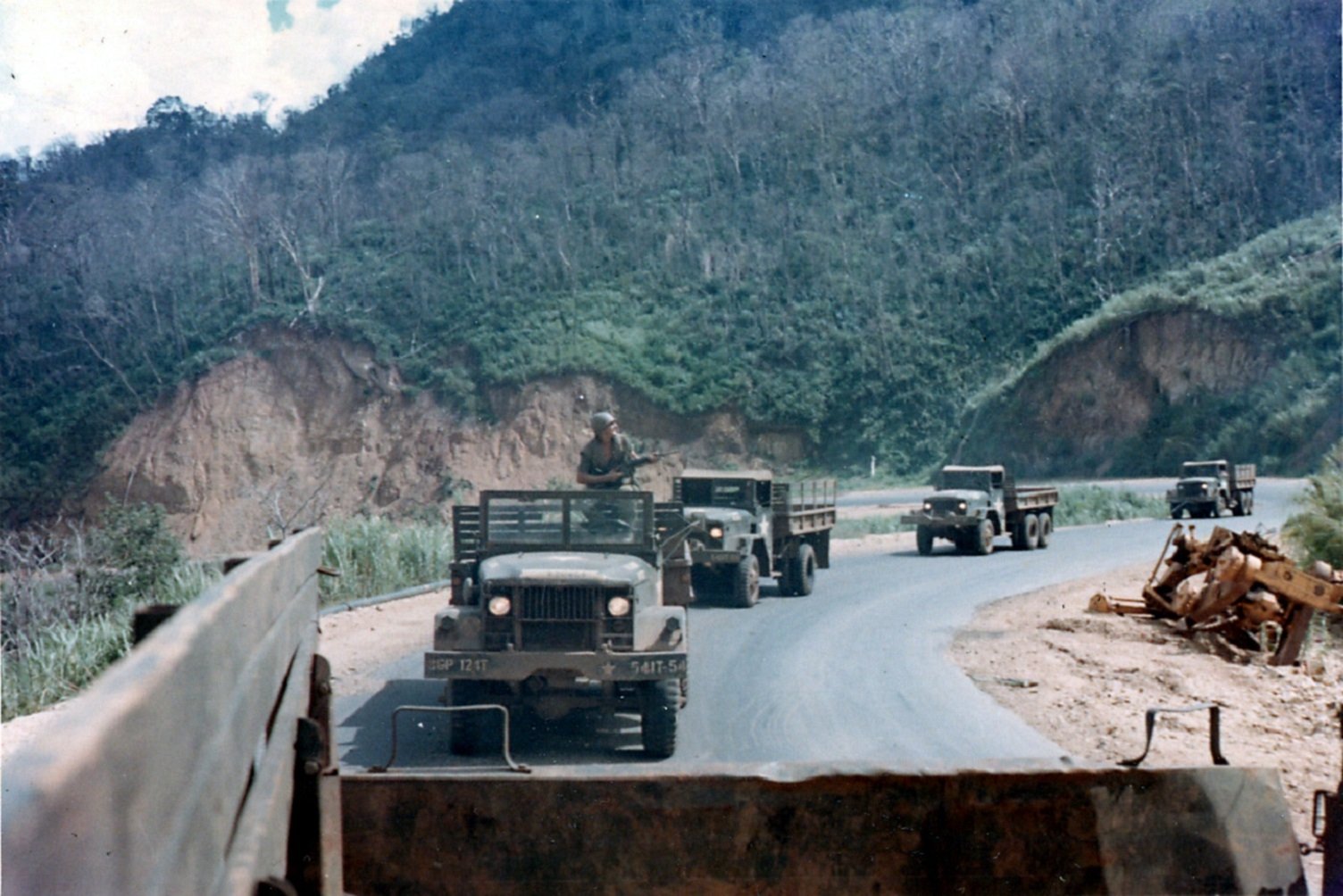 HD desktop wallpaper showing military trucks convoy on a winding road during the Vietnam War, set against lush green hills and rugged terrain.