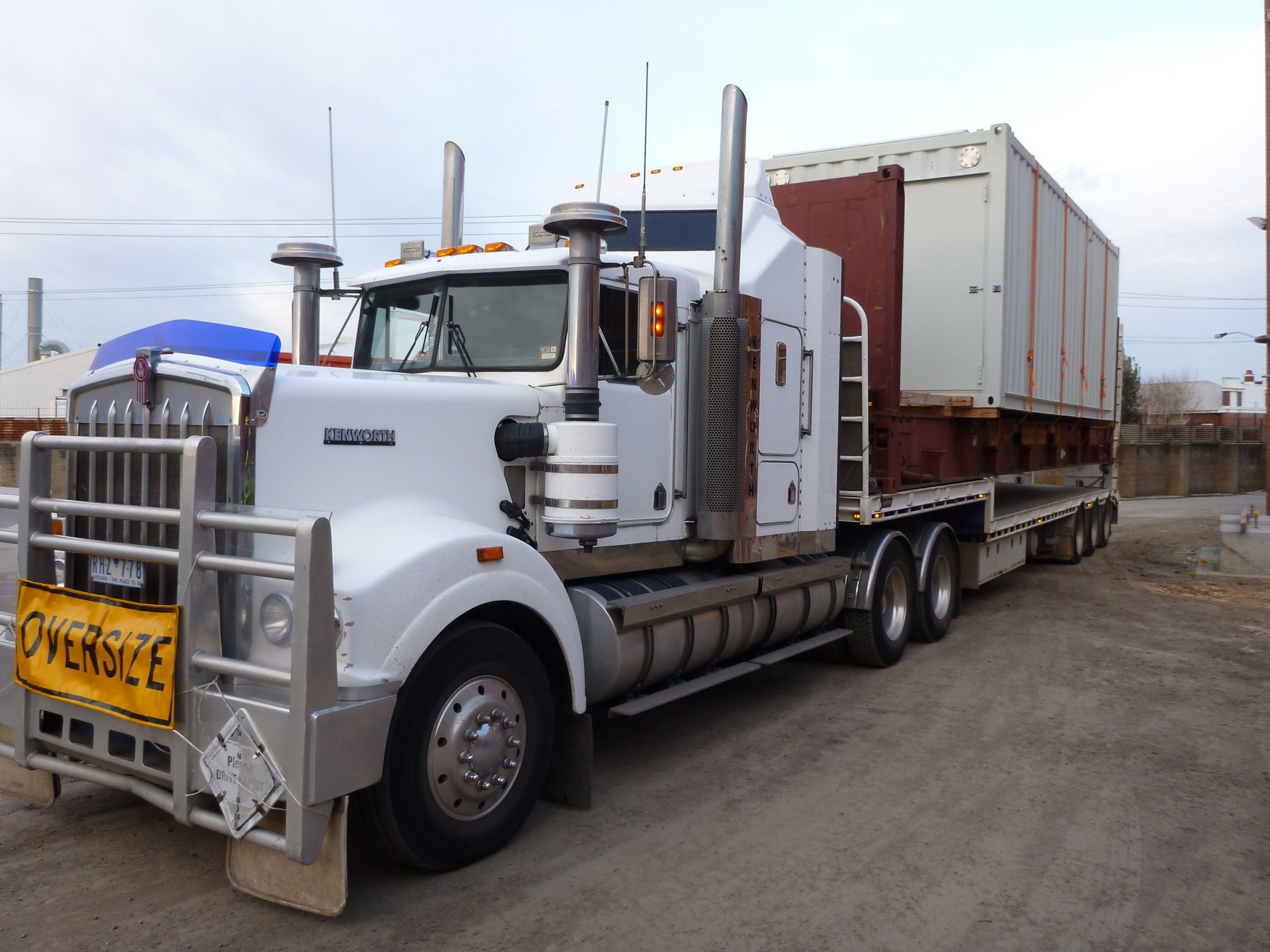 Kenworth vehicle: white semi truck with flatbed carrying oversized cargo in an industrial yard — HD PC desktop wallpaper background.