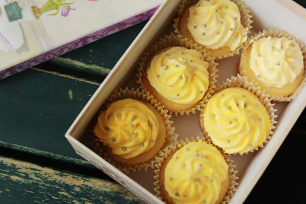HD PC desktop wallpaper: box of yellow-iced cupcakes with swirled icing and silver sprinkles on a rustic table — dessert sweets, cake-style food background.