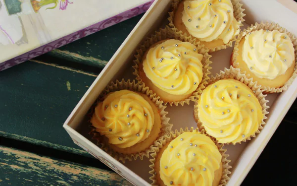 HD PC desktop wallpaper: box of yellow-iced cupcakes with swirled icing and silver sprinkles on a rustic table — dessert sweets, cake-style food background.