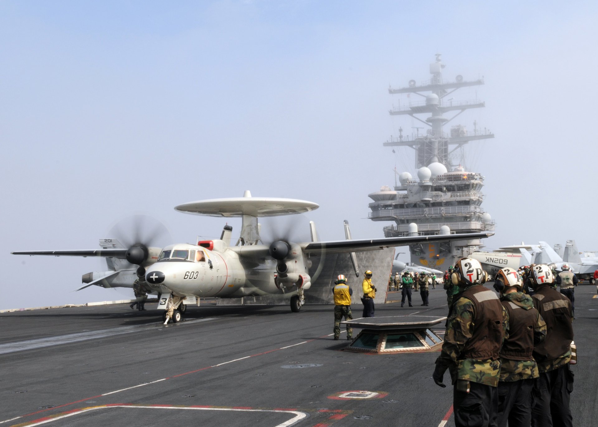 USS Ronald Reagan (CVN-76) aircraft carrier deck with a Northrop Grumman E-2 Hawkeye and crew members preparing for flight operations.