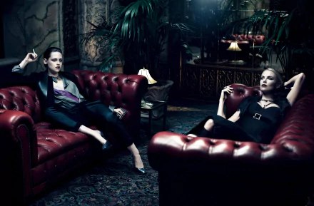 A stylish HD wallpaper featuring Charlize Theron and Kristen Stewart lounging on red leather couches in a vintage, lush setting, exuding an air of elegance and celebrity allure.