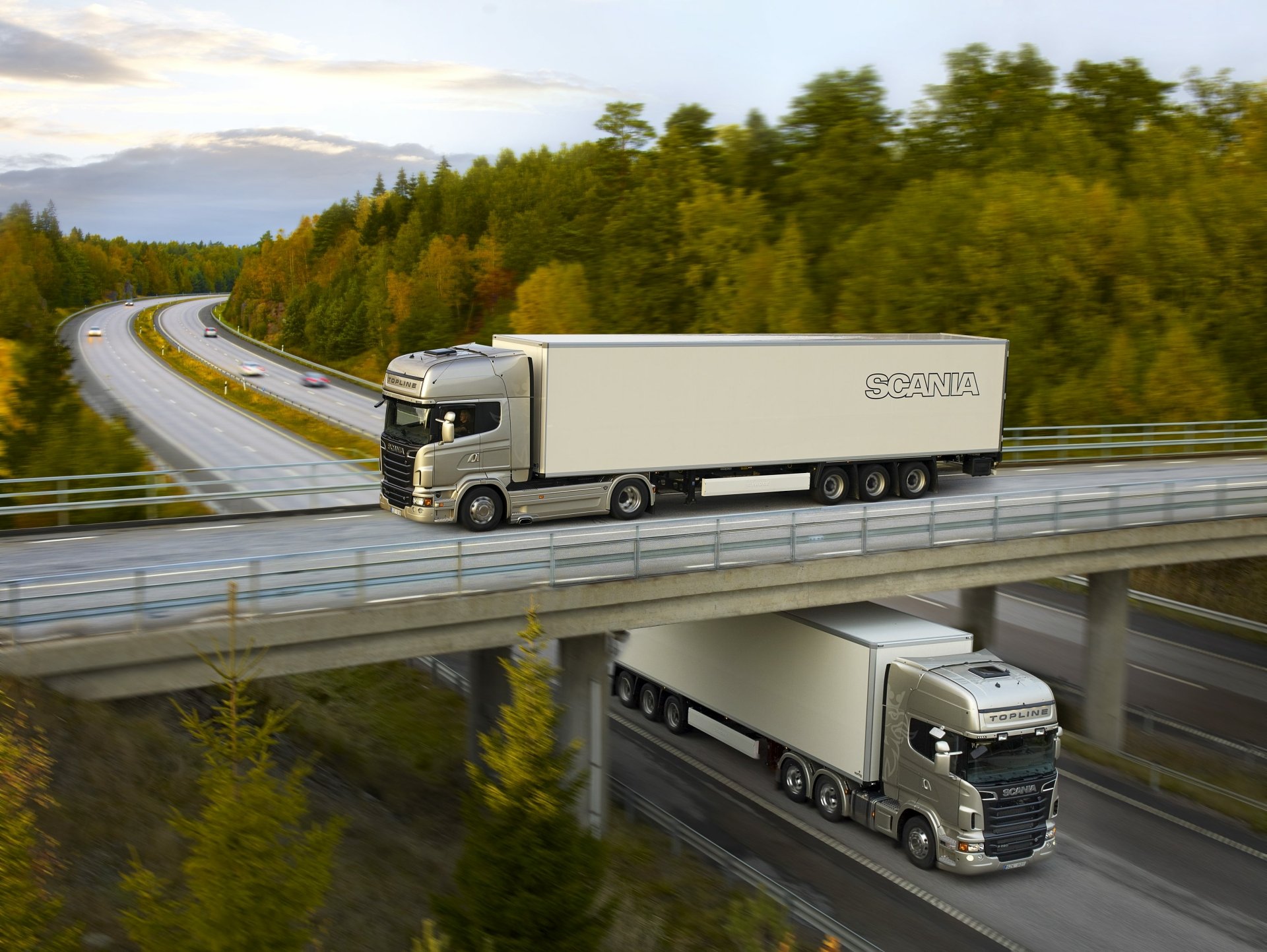 2K Quad HD PC desktop wallpaper background: two Scania trucks crossing a highway overpass through a forested landscape at dusk.