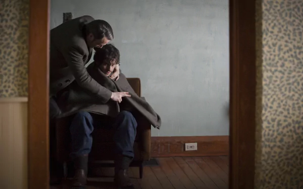 Moody TV-drama scene: one man leans over another seated in a sparse, dim room; atmospheric 2K Quad HD PC desktop wallpaper/background.