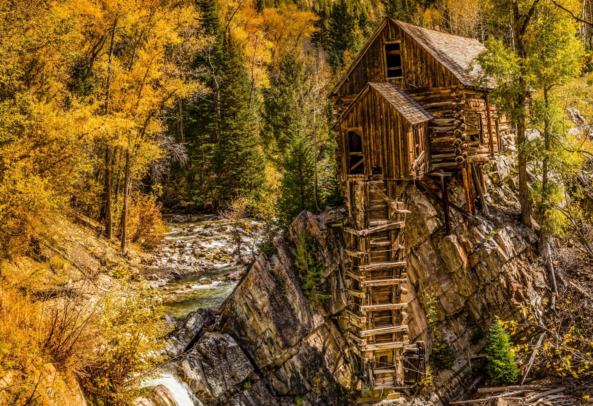 HDR 4K Ultra HD desktop wallpaper of man-made Crystal Mill perched on a rocky ledge above a rushing stream, surrounded by golden autumn trees.