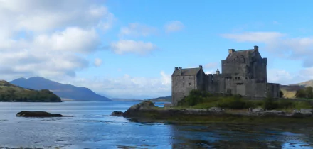 2K Quad HD PC desktop wallpaper: man-made Eilean Donan Castle on a rocky promontory overlooking calm sea and distant hills beneath a blue sky.