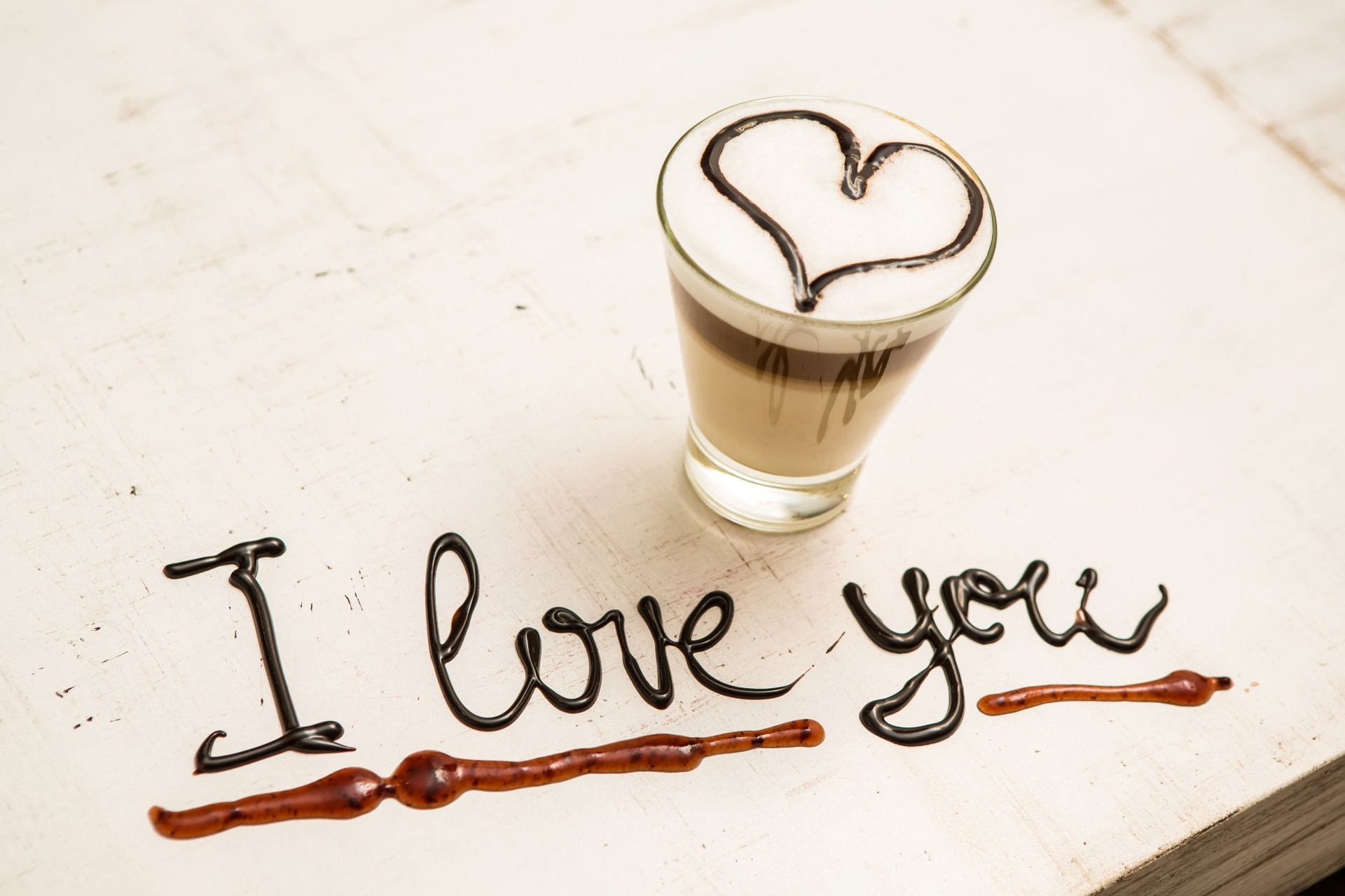 A romantic scene featuring a coffee glass adorned with chocolate, elegantly forming a heart and the words I love you, capturing love and indulgence in a warm, inviting atmosphere.