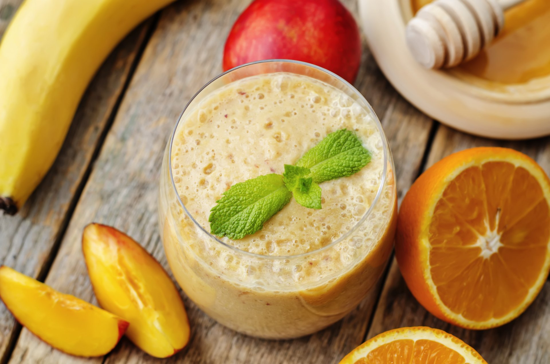 A vibrant 4K Ultra HD wallpaper featuring a creamy smoothie garnished with mint, surrounded by fresh orange slices, banana, peach, and honey on a rustic wooden surface.