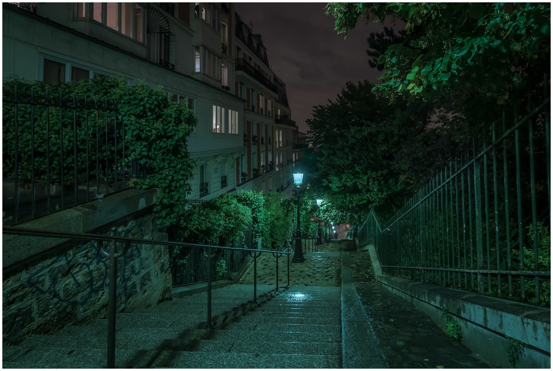 Night view of a quiet Montmartre street in Paris, featuring stone stairs illuminated by a glowing lamp post surrounded by greenery, captured in 4K Ultra HD.