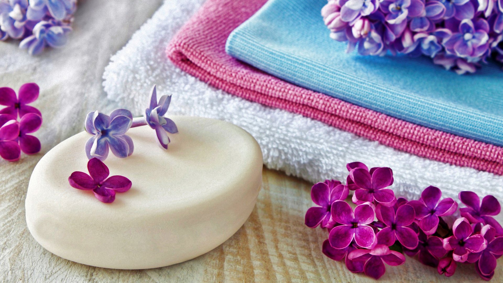 Close-up of lilac flowers beside a smooth bar of soap and soft towels, creating a serene spa atmosphere. This HD wallpaper captures a tranquil and rejuvenating vibe.