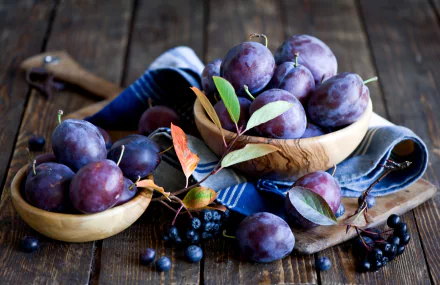 HD PC desktop wallpaper featuring ripe purple plums in wooden bowls on a rustic wooden table with scattered leaves and berries, highlighting fresh fruit and food themes.