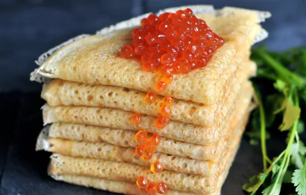 Stack of folded crêpes topped with bright orange caviar, set against a dark background with green herbs, captured in HD for a vibrant desktop wallpaper.