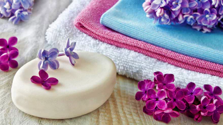 Close-up of lilac flowers beside a smooth bar of soap and soft towels, creating a serene spa atmosphere. This HD wallpaper captures a tranquil and rejuvenating vibe.