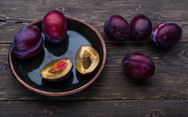 Ripe plums and a halved plum with exposed pit on a ceramic plate and dark wooden table — fruit/food image as a 5K Ultra HD PC desktop wallpaper background.