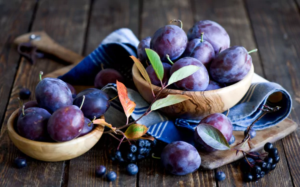 HD PC desktop wallpaper featuring ripe purple plums in wooden bowls on a rustic wooden table with scattered leaves and berries, highlighting fresh fruit and food themes.