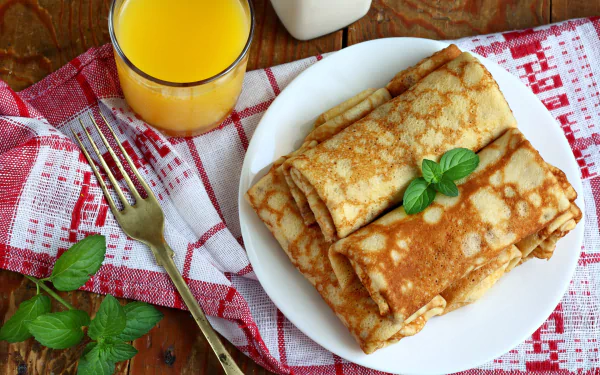 A delicious plate of crêpes garnished with fresh mint, accompanied by a glass of juice, all set on a rustic wooden table with a red checkered cloth.