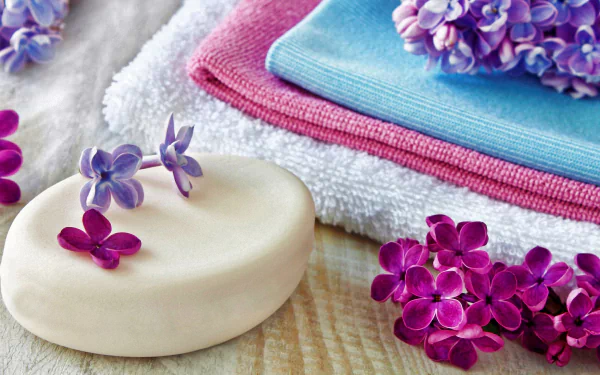 Close-up of lilac flowers beside a smooth bar of soap and soft towels, creating a serene spa atmosphere. This HD wallpaper captures a tranquil and rejuvenating vibe.