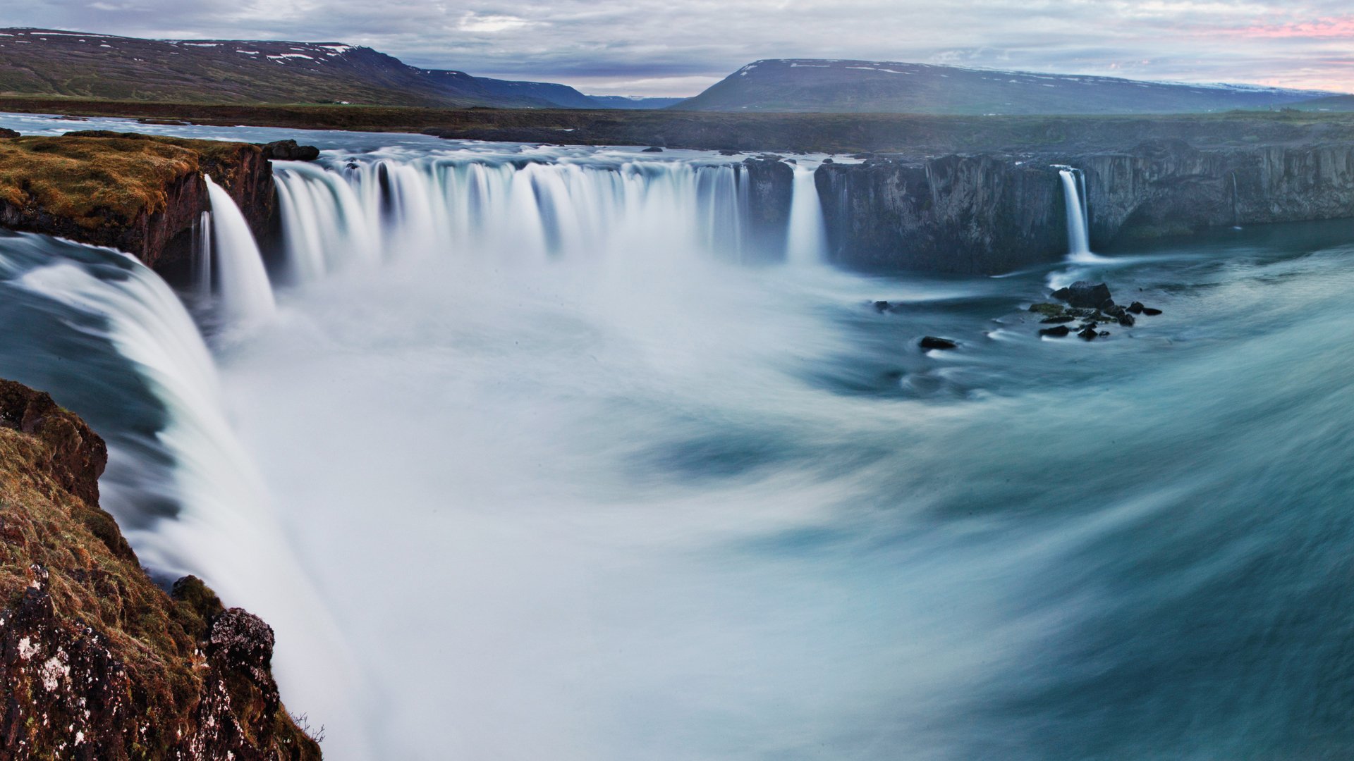A stunning HD wallpaper featuring a majestic waterfall cascading over rugged cliffs, surrounded by breathtaking mountain landscapes and tranquil waters.